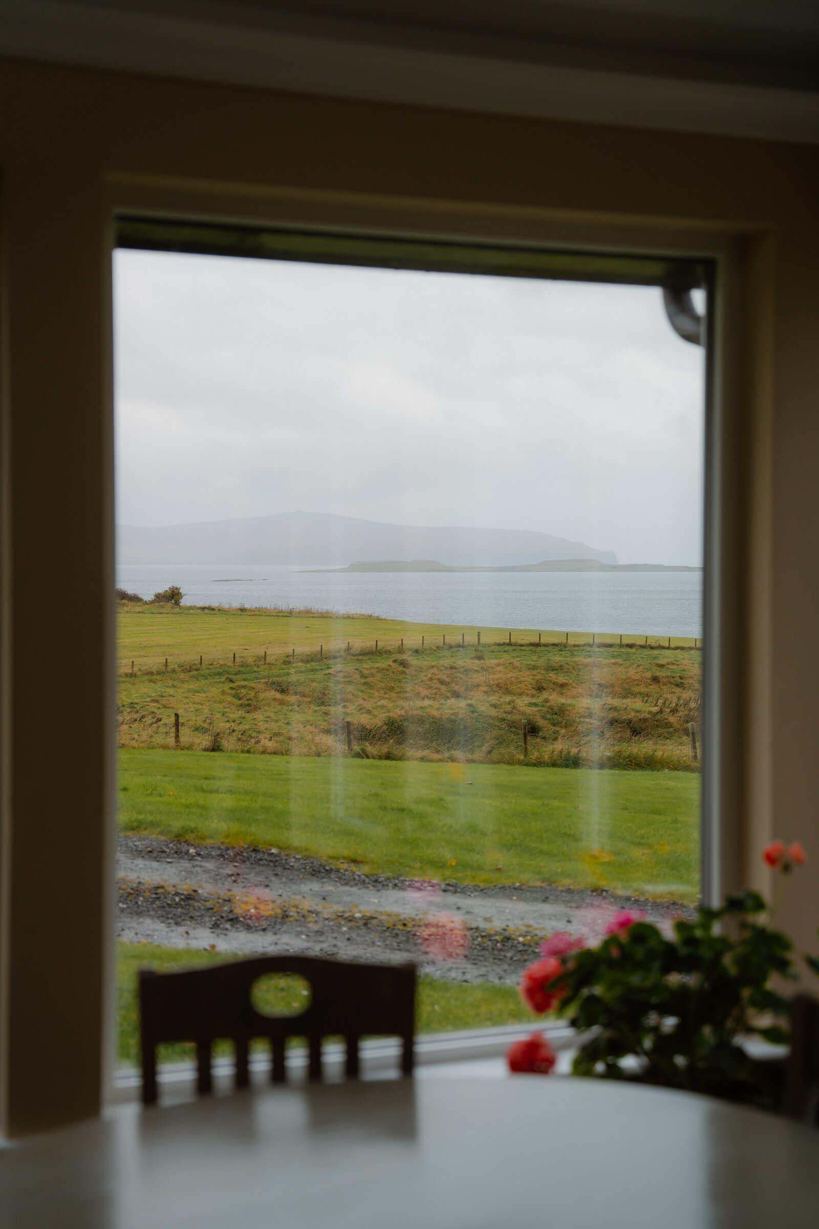 A dining chair and potted flowers sit before a window looking out over fields to the sea and distant islands.