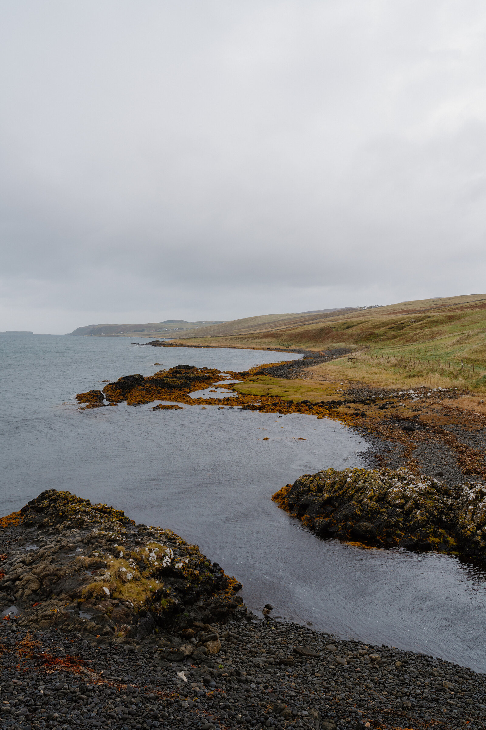 Rocky, lichen-covered outcrops edge a calm bay beneath rolling green hills.