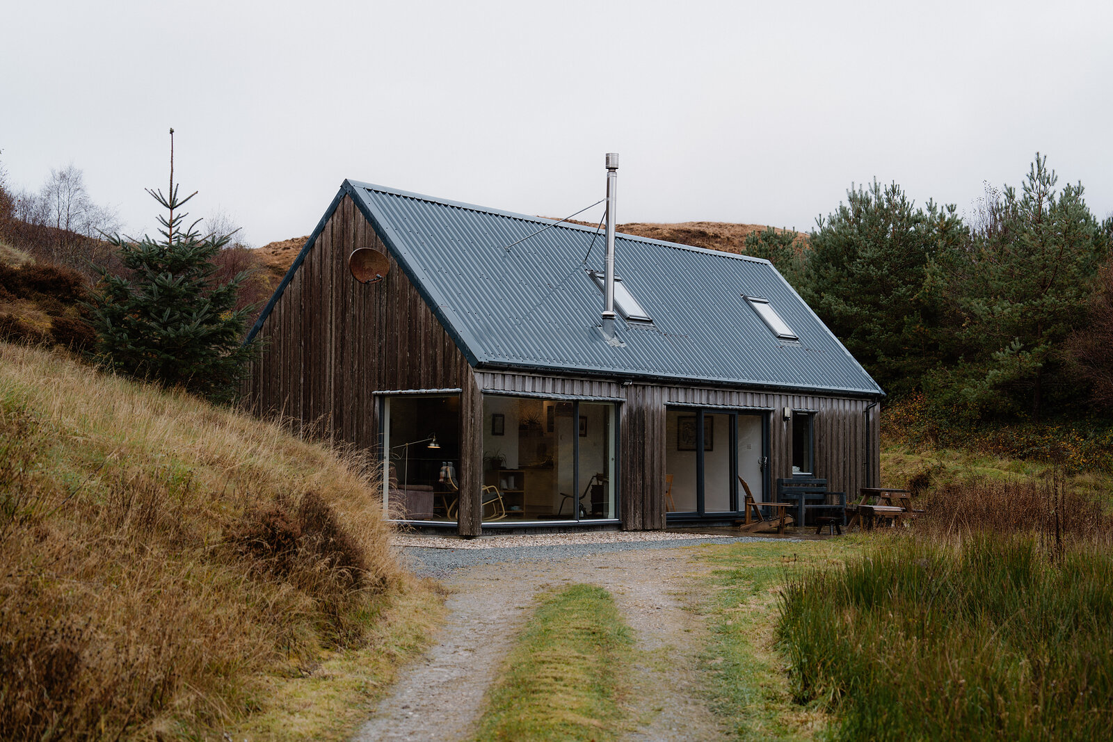 A dark timber-clad cabin with a corrugated metal roof sits on a gravel drive amid moorland scrub.