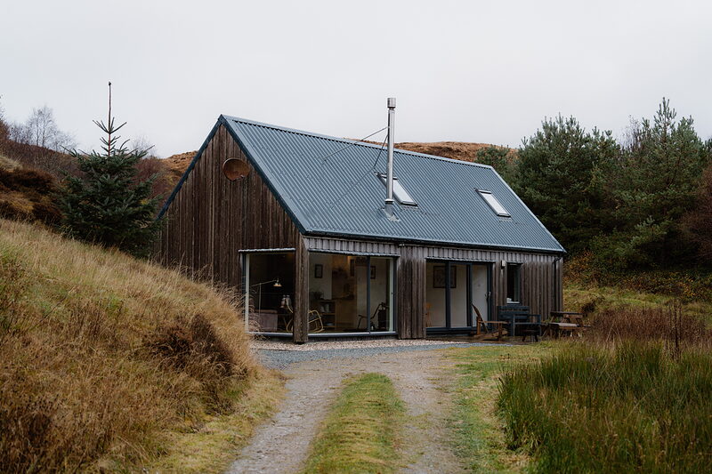 A dark timber-clad cabin with a corrugated metal roof sits on a gravel drive amid moorland scrub.