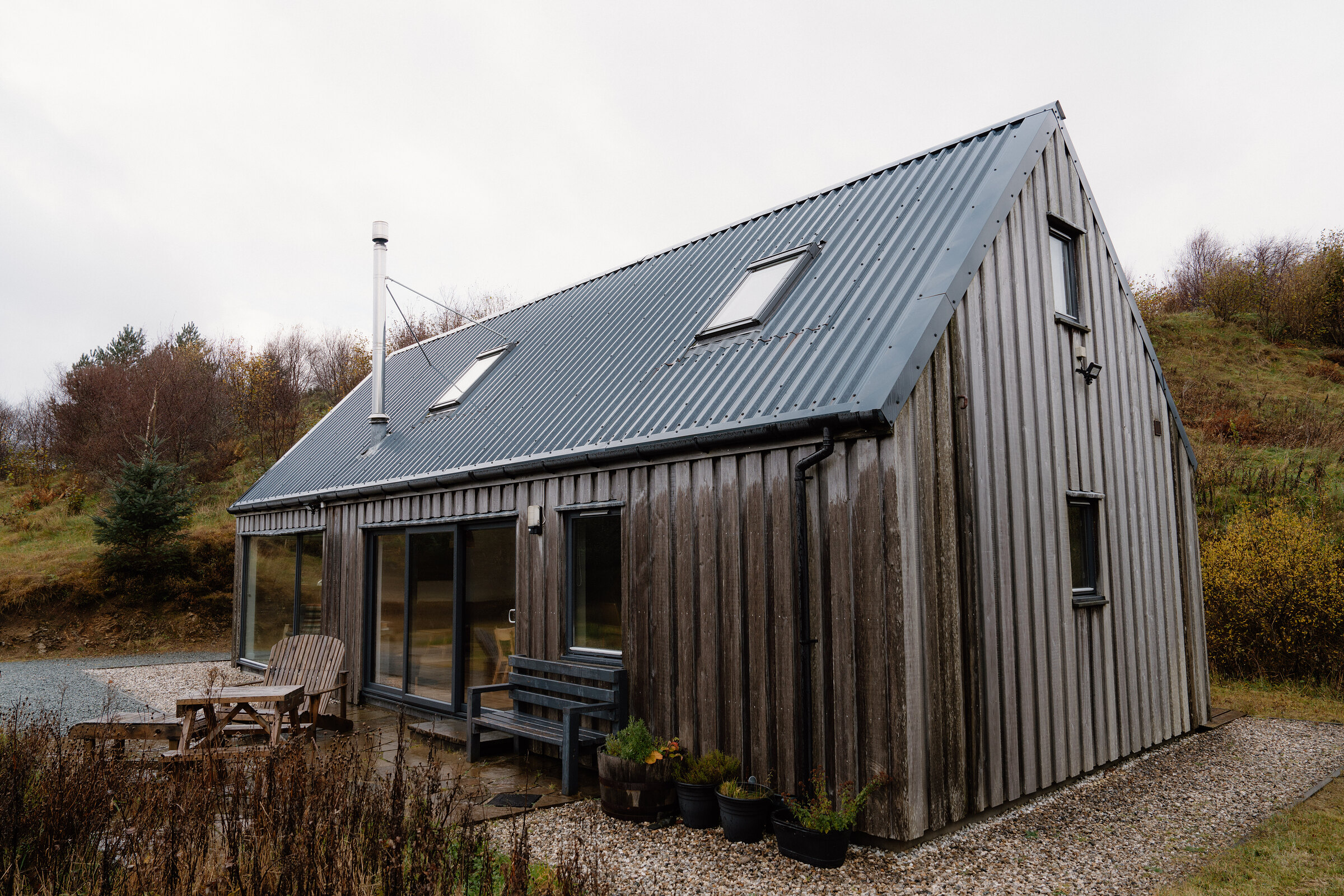 Stonechat Bothy from the side, its tall gable and roof lights, with a picnic table and bench on the gravel and moorland behind.