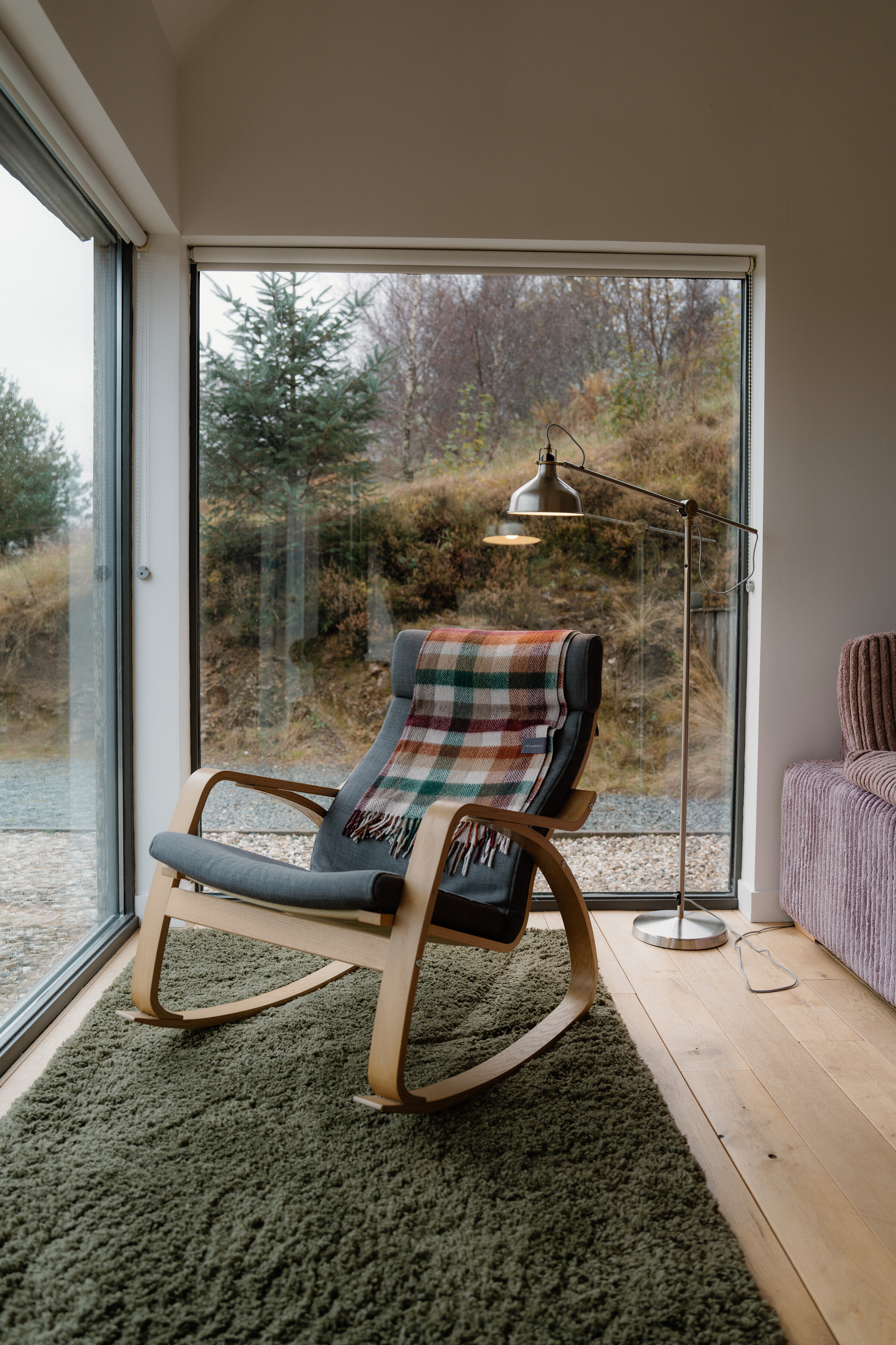 A rocking chair with a tartan cushion on a green rug in the corner, floor-to-ceiling windows onto the birches and the track.