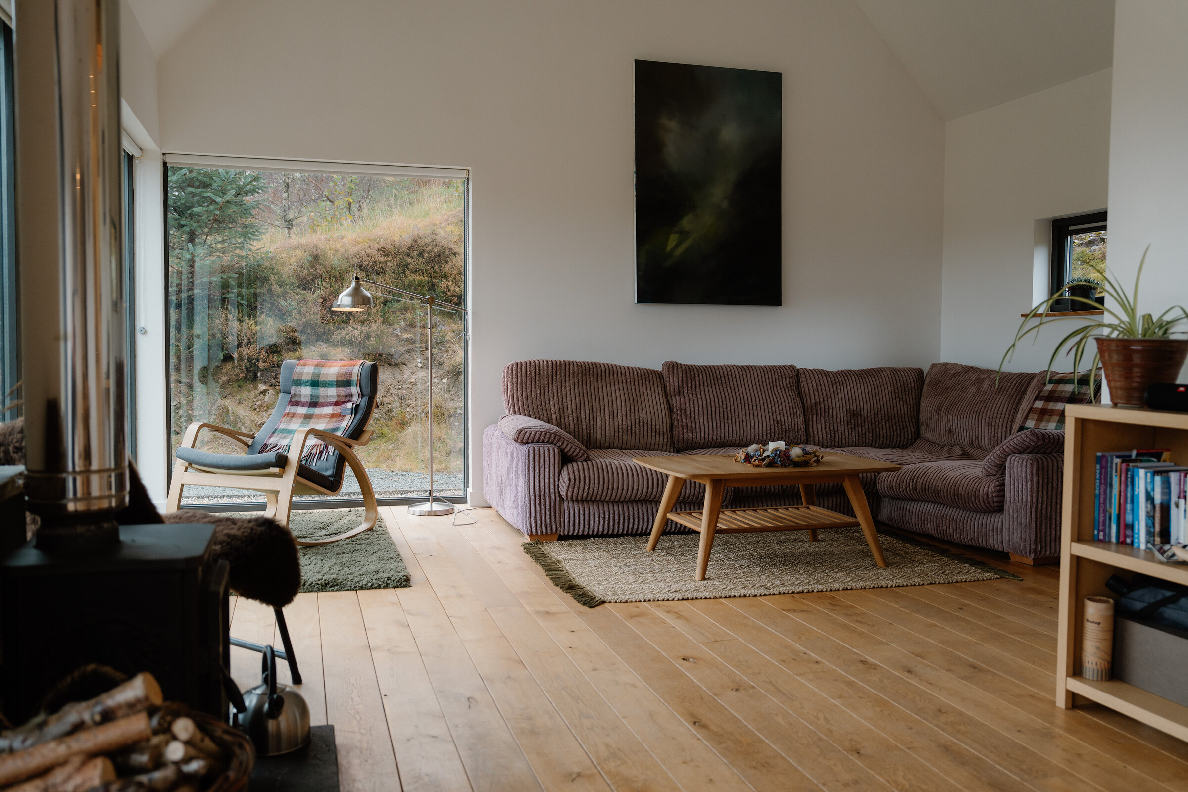 The sitting room: a corduroy sofa, a rocking chair by the corner windows, a bookcase and the wood stove with its log basket.