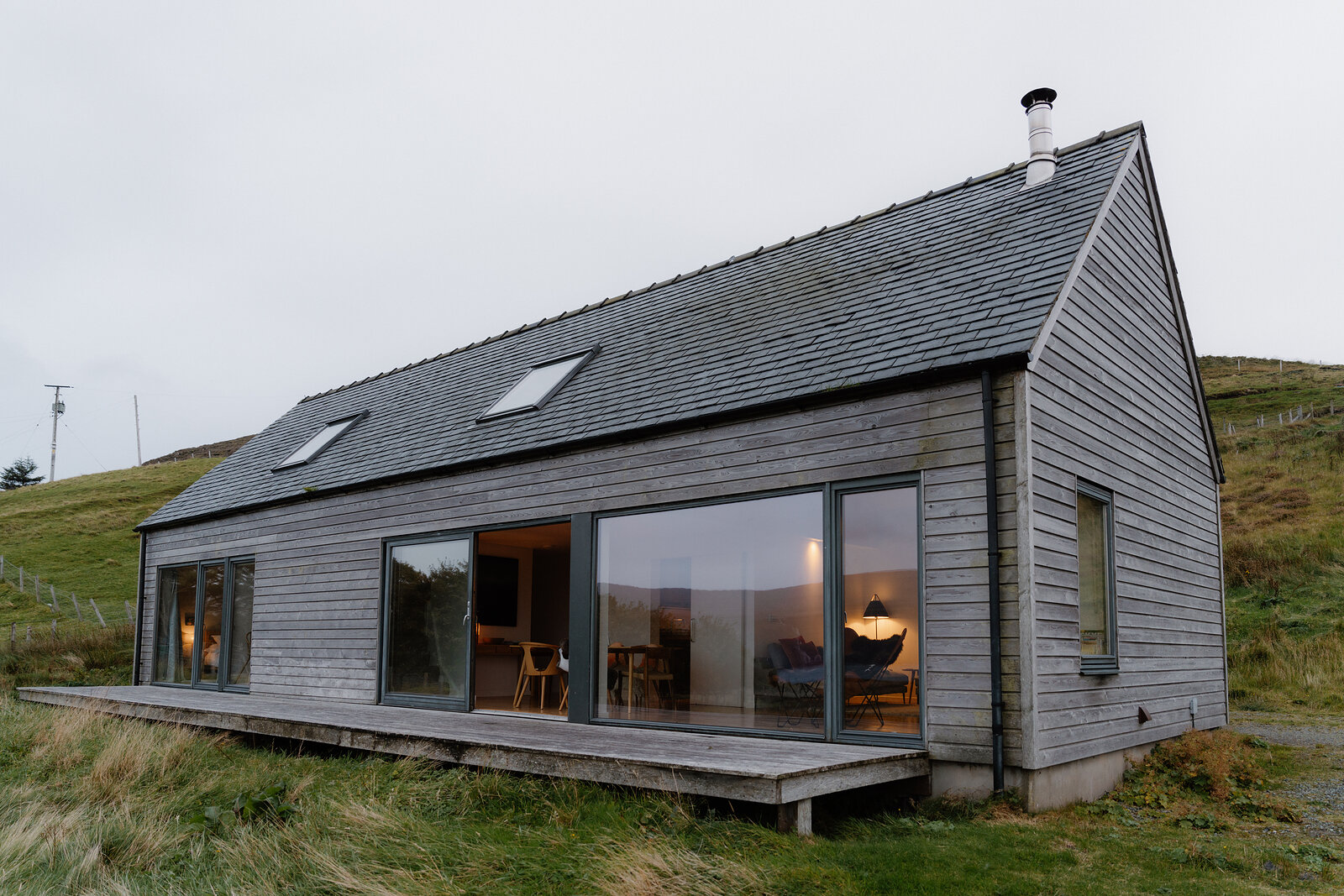 A dark timber-clad gabled cabin with a chimney and skylights glows warmly from within at dusk on a grassy hillside.