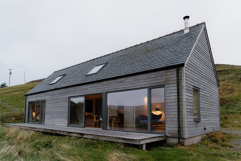 A dark timber-clad gabled cabin with a chimney and skylights glows warmly from within at dusk on a grassy hillside.