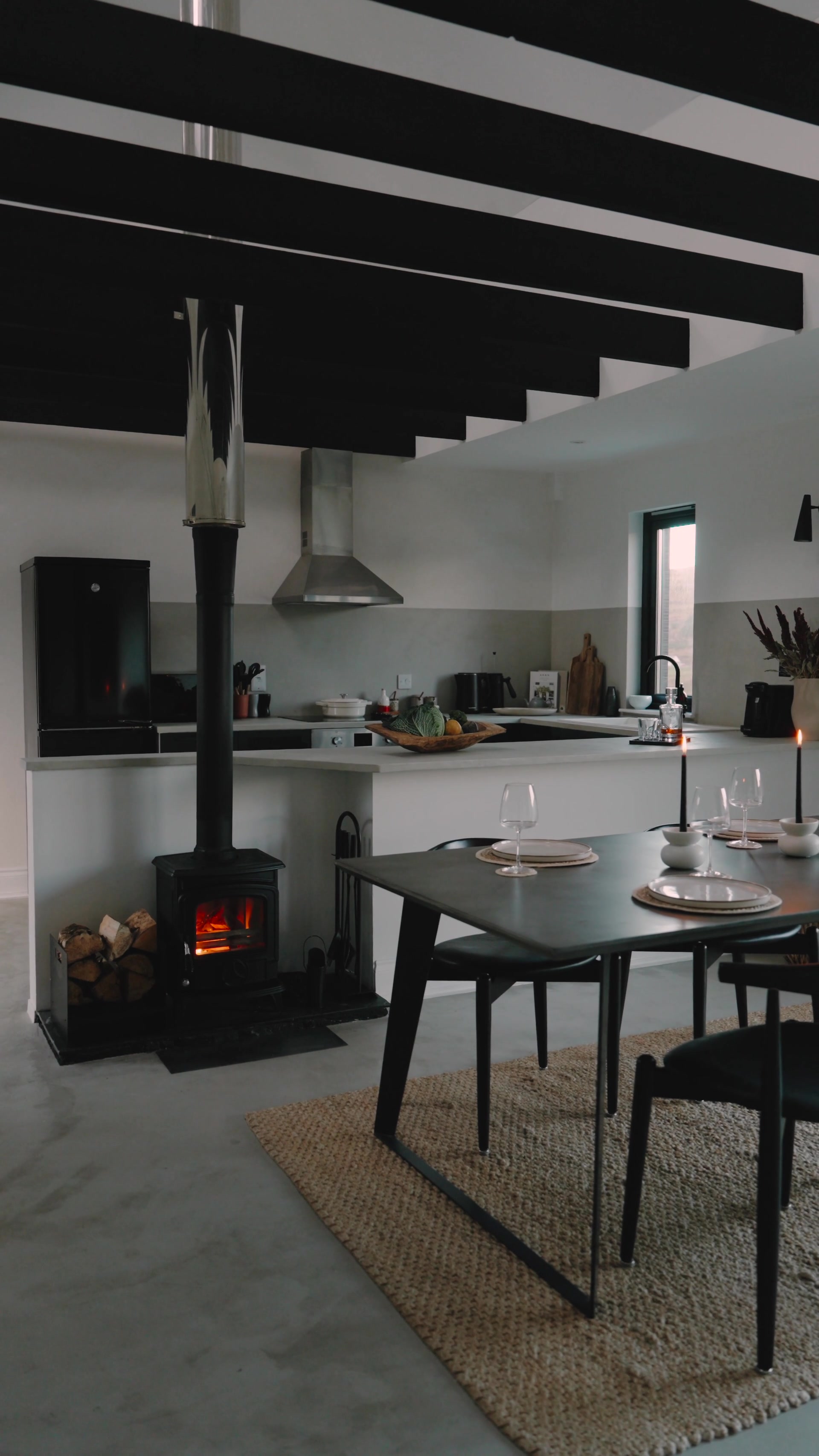 A wood-burning stove glows beside a black dining table set with wine glasses, candles and plates, an open-plan kitchen with a stainless steel extractor hood behind.