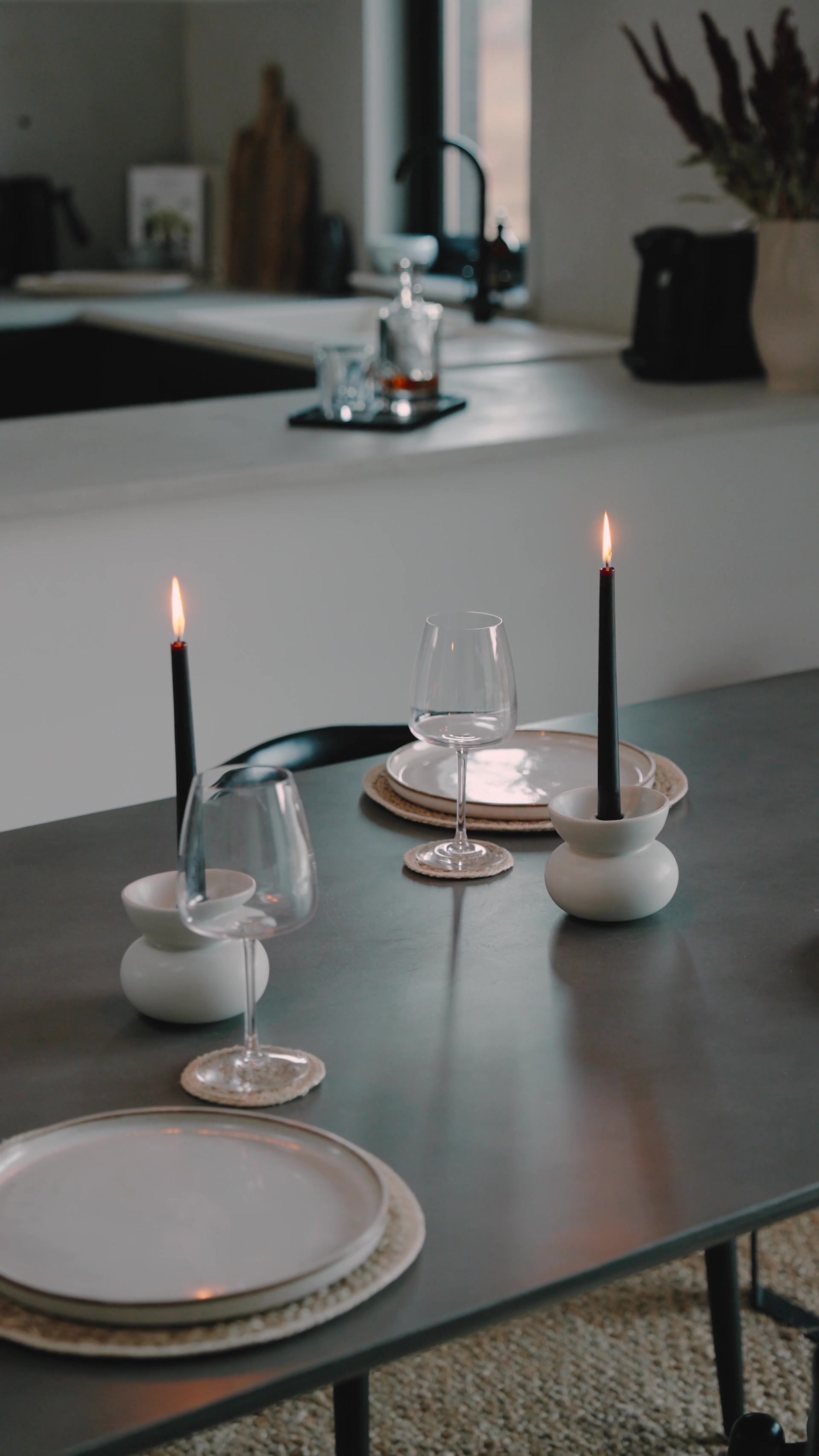 Two lit black taper candles in round white holders flank stemmed wine glasses on a dark dining table, a whisky decanter and tumbler set on a tray by the kitchen sink behind.