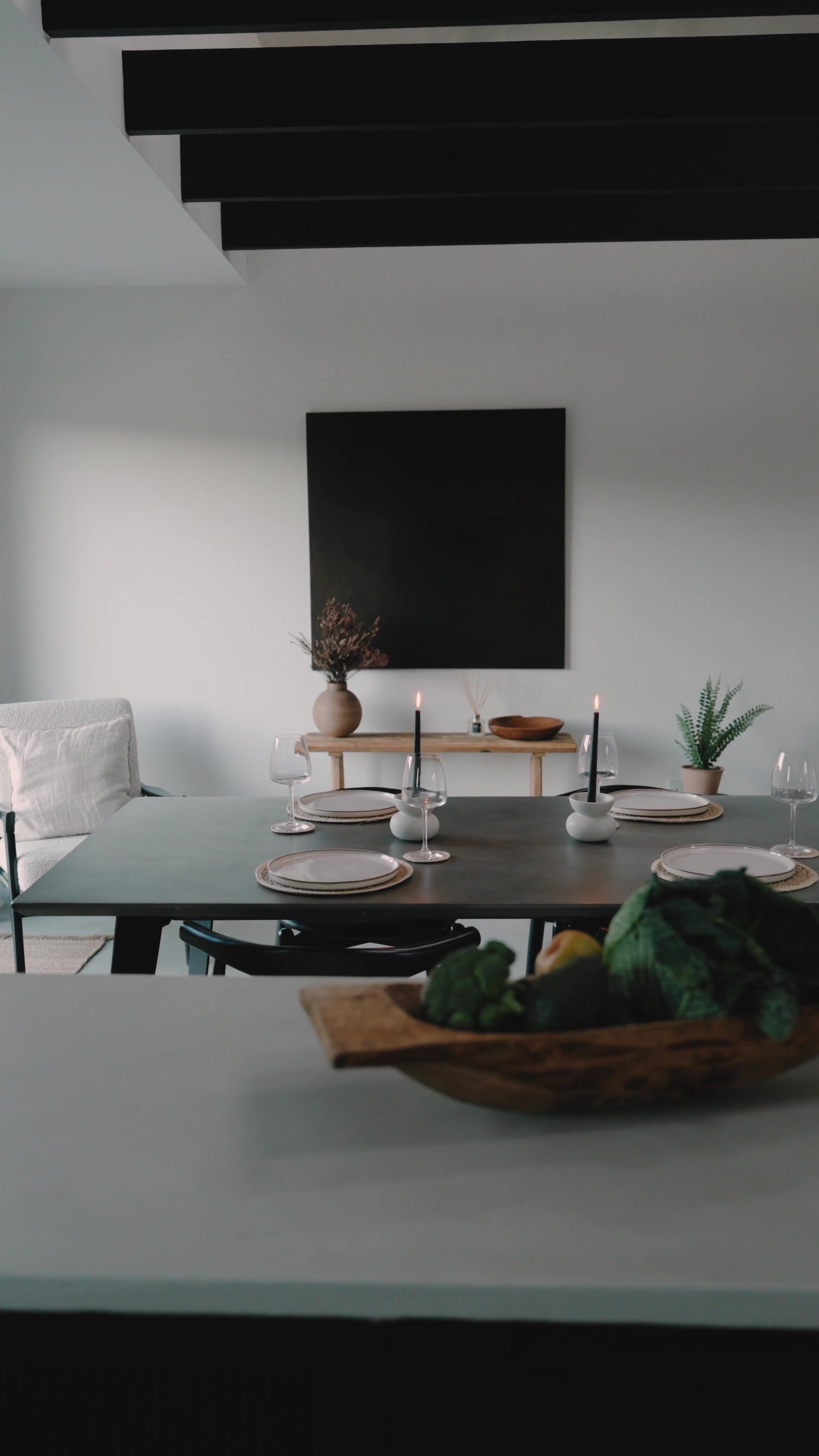 A dining table set with white plates, wine glasses and lit black taper candles stands before a large black square artwork, a wooden bowl of vegetables in the foreground.