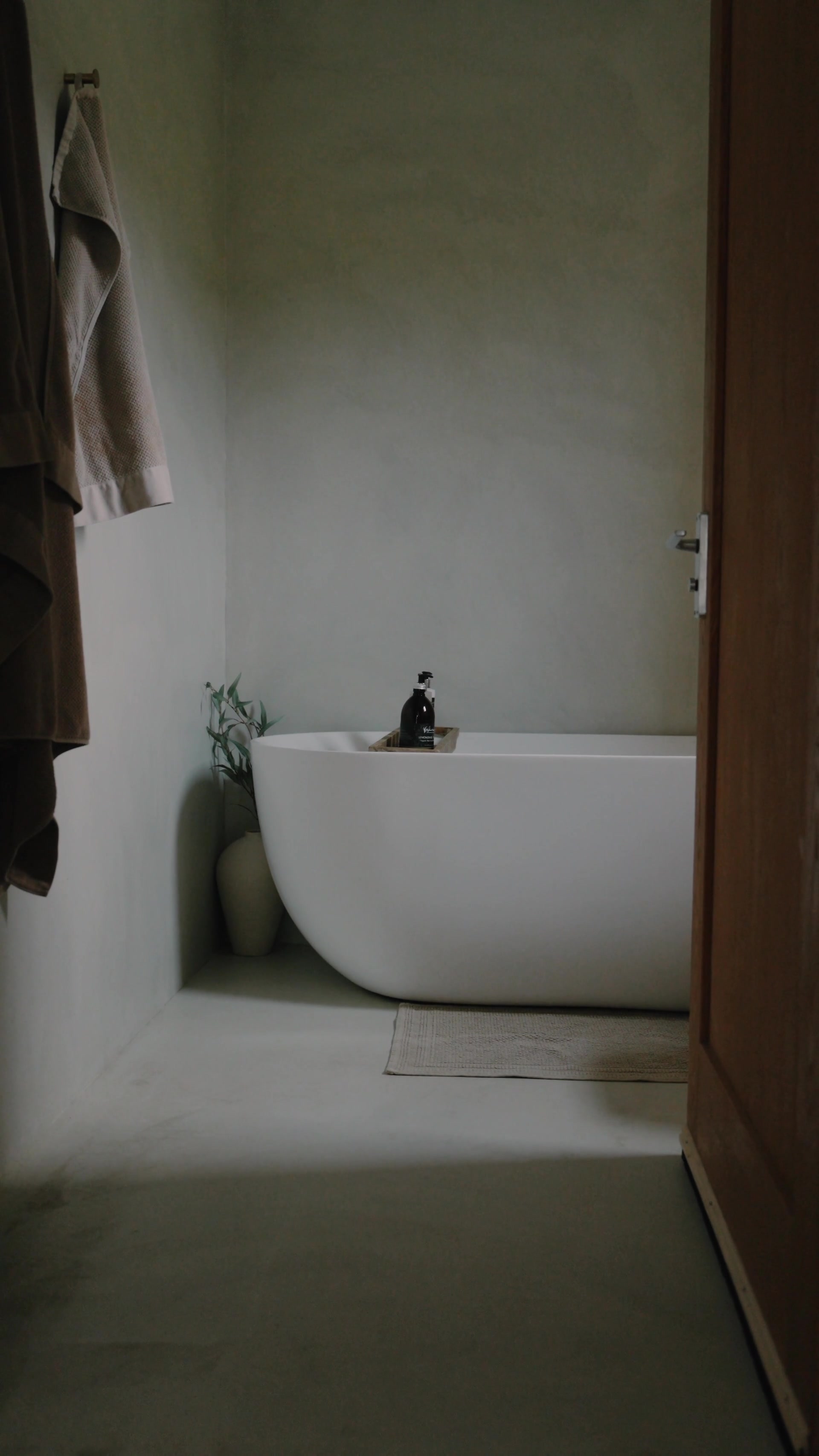 A freestanding white oval bathtub sits in the corner of a plastered bathroom, a striped towel hanging on a hook and a small vase of foliage beside it.