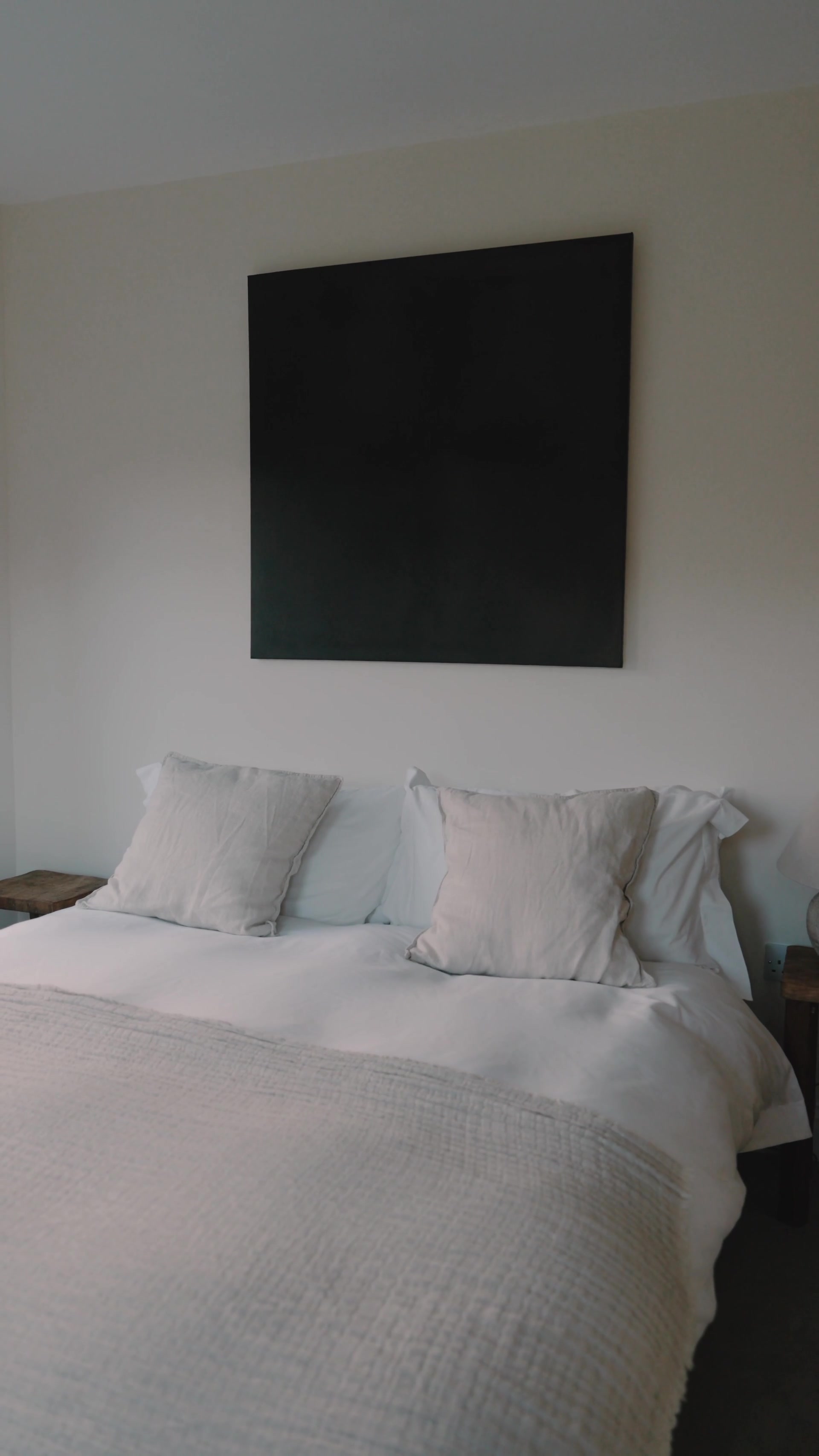 A large black square canvas hangs above a bed dressed in white linen and pale cushions, a woven throw folded across the foot of the mattress.