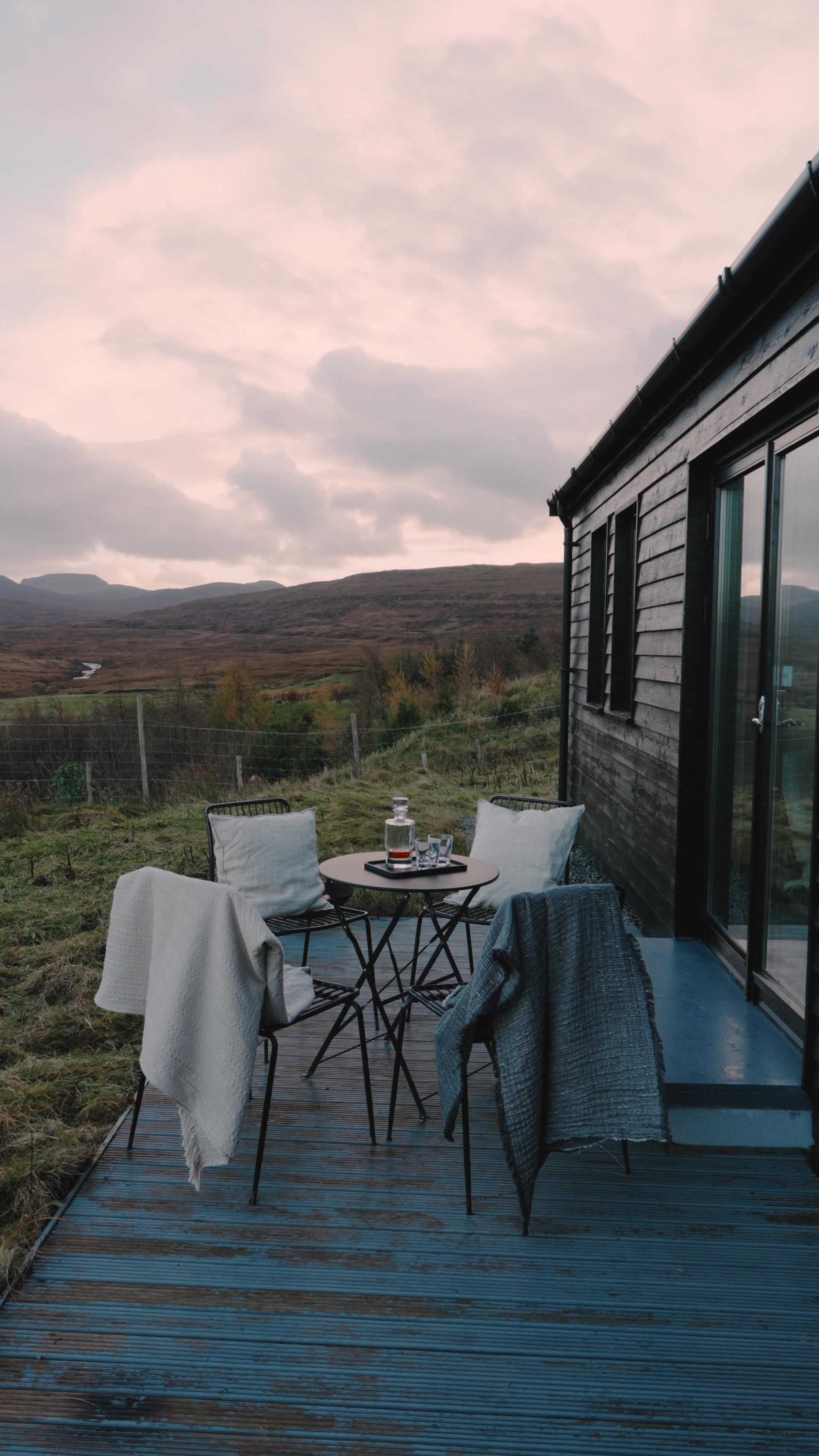 Two metal bistro chairs draped with blankets flank a small round table set with a whisky decanter and glasses on a wooden deck, moorland and hills stretching beyond at dusk.