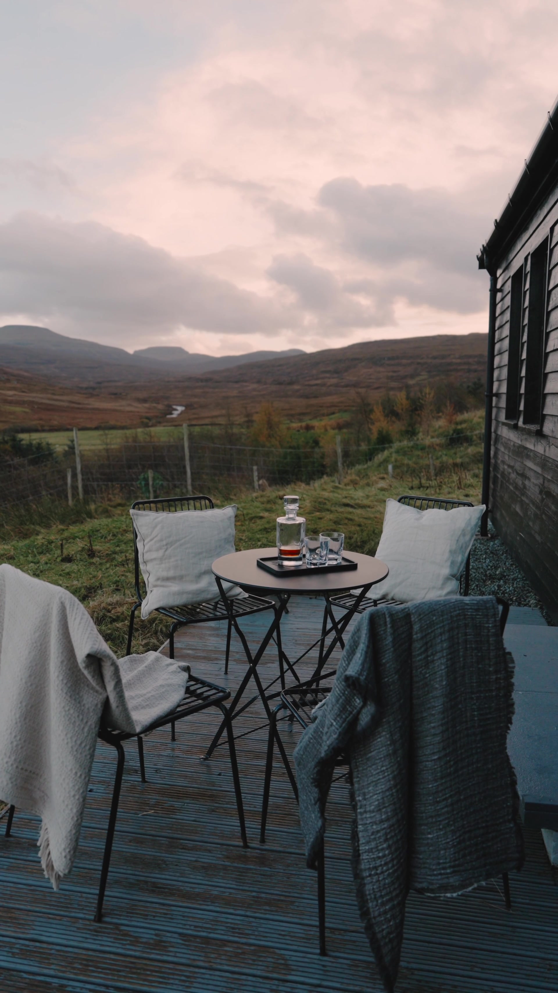 A glass whisky decanter and two tumblers sit on a tray on a small round table between two cushioned chairs draped with blankets, moorland hills glowing pink at dusk beyond a timber cabin.