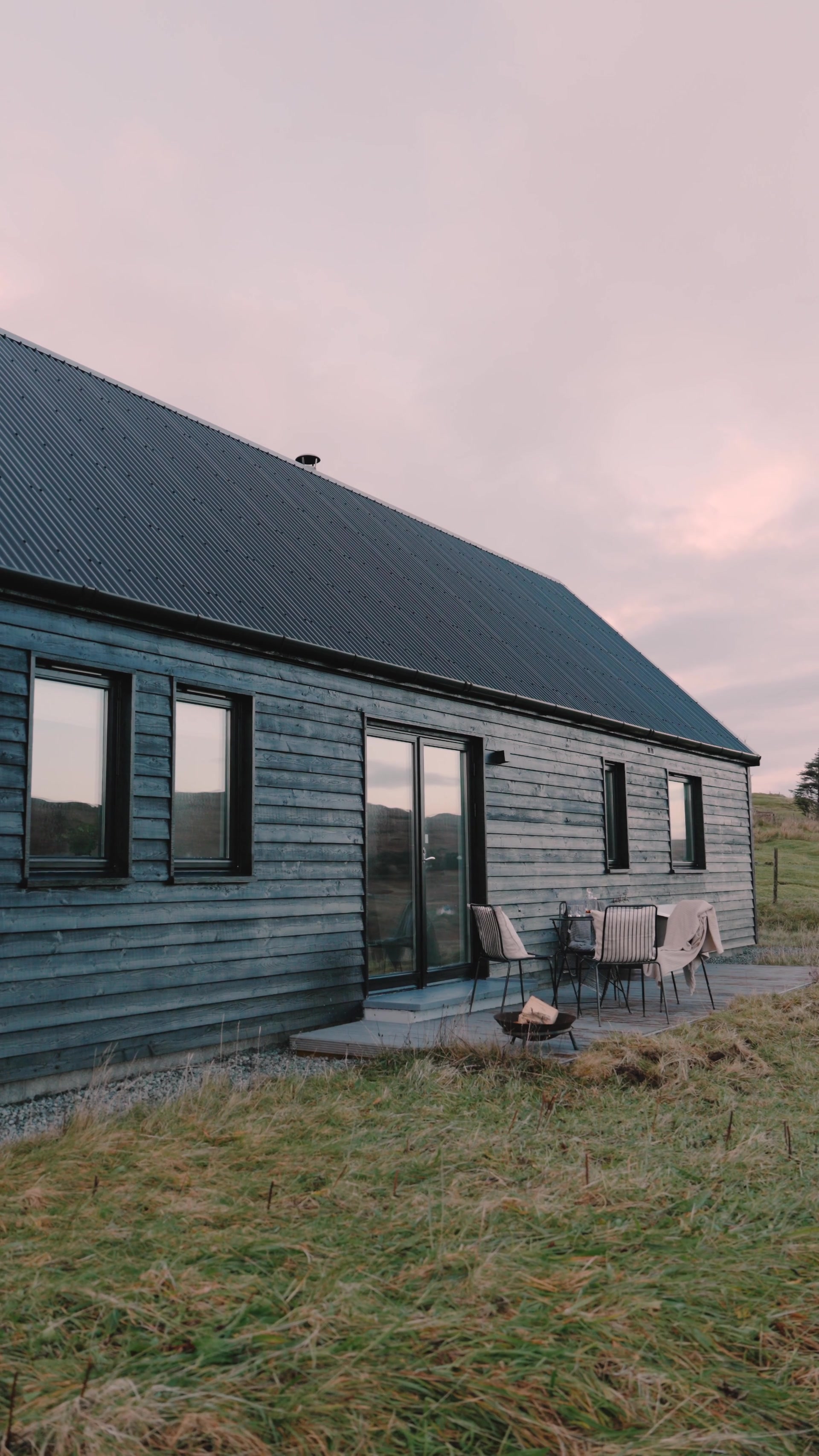 A dark timber cabin with skylight windows and a glazed sliding door stands on a gravel path, a small outdoor dining set on the deck beside it under a pink dusk sky.