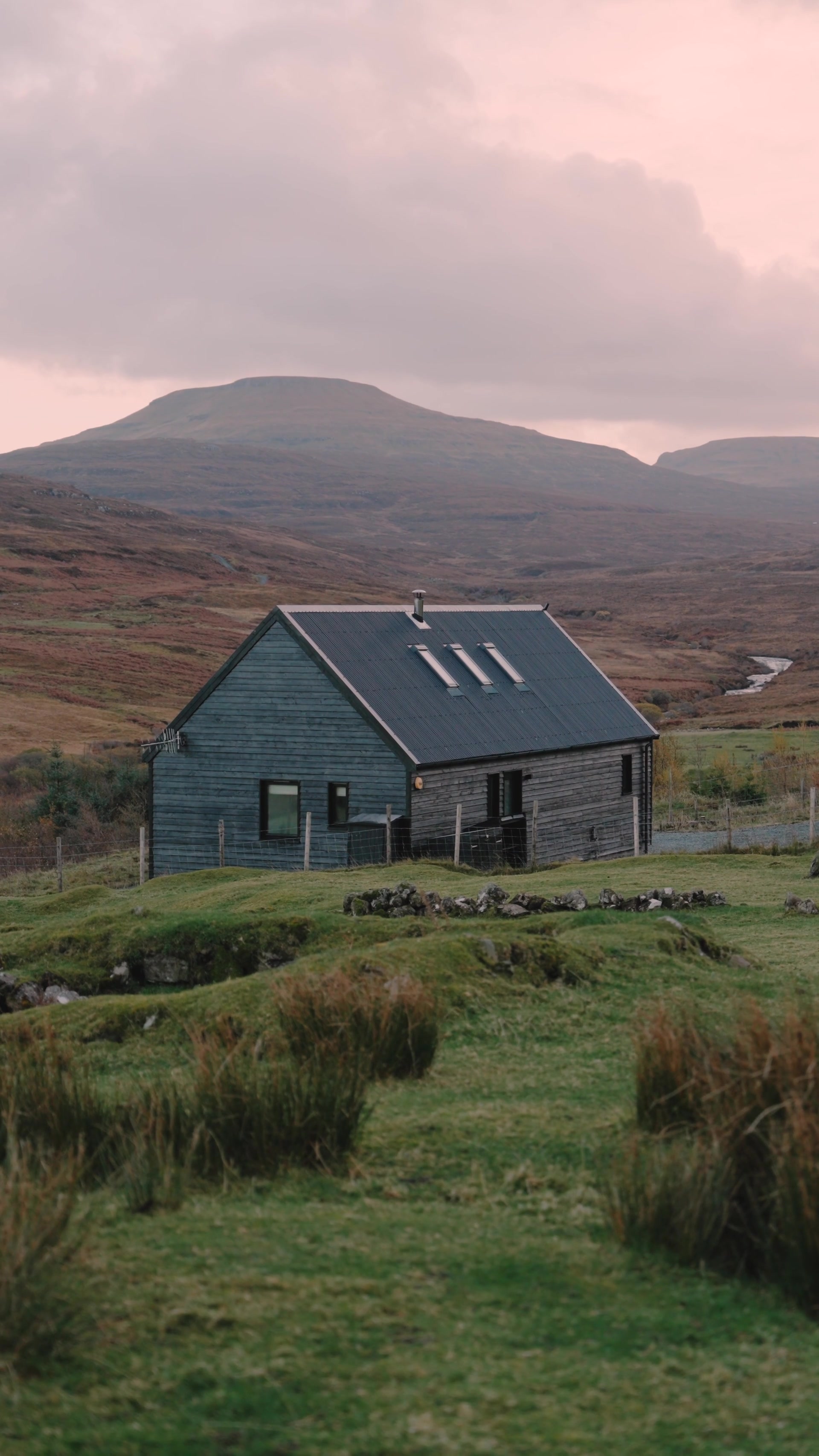 A dark timber cabin with a pitched corrugated roof sits alone in open moorland, a river winding below distant hills under a soft pink evening sky.