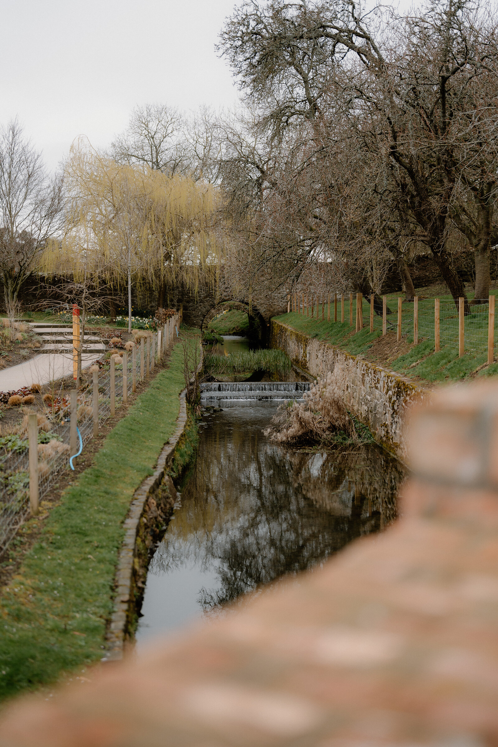 A small stone bridge crosses a weir on a stream running through gardens at Glenkinchie.