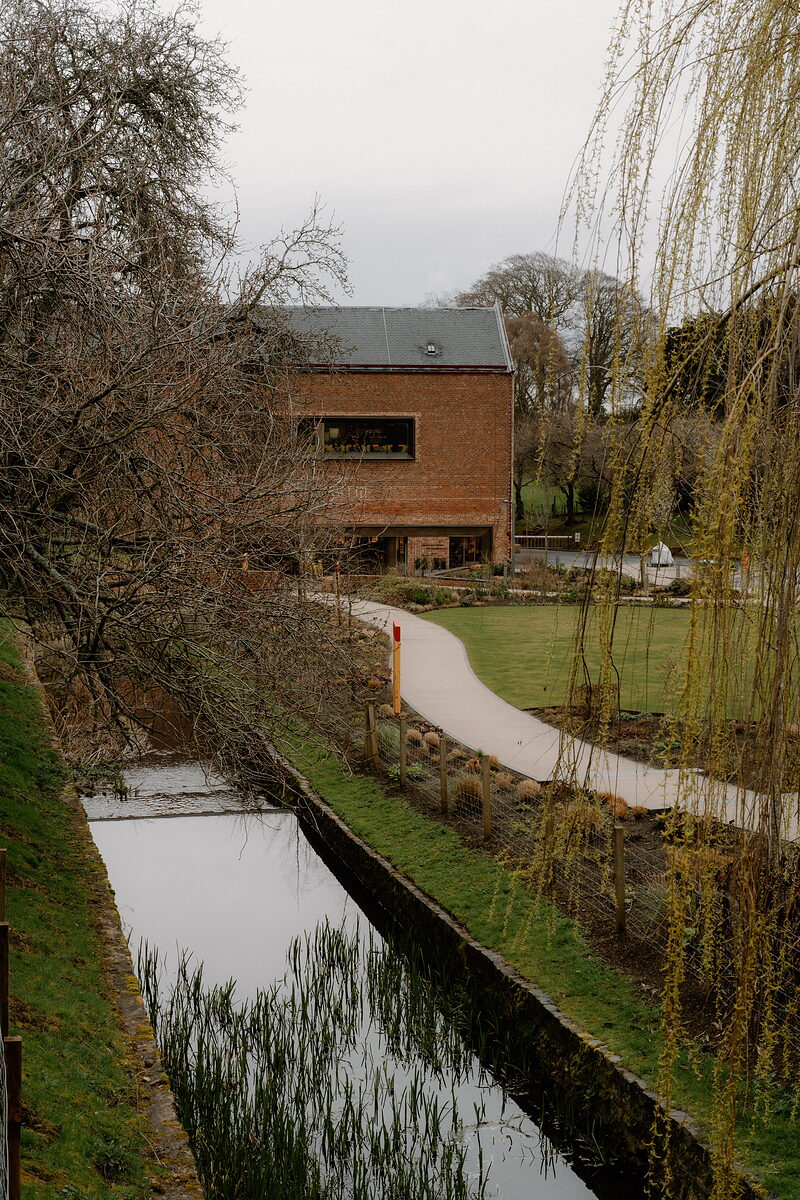 The red-brick Glenkinchie building overlooks a garden path beside a stream lined with willow trees.