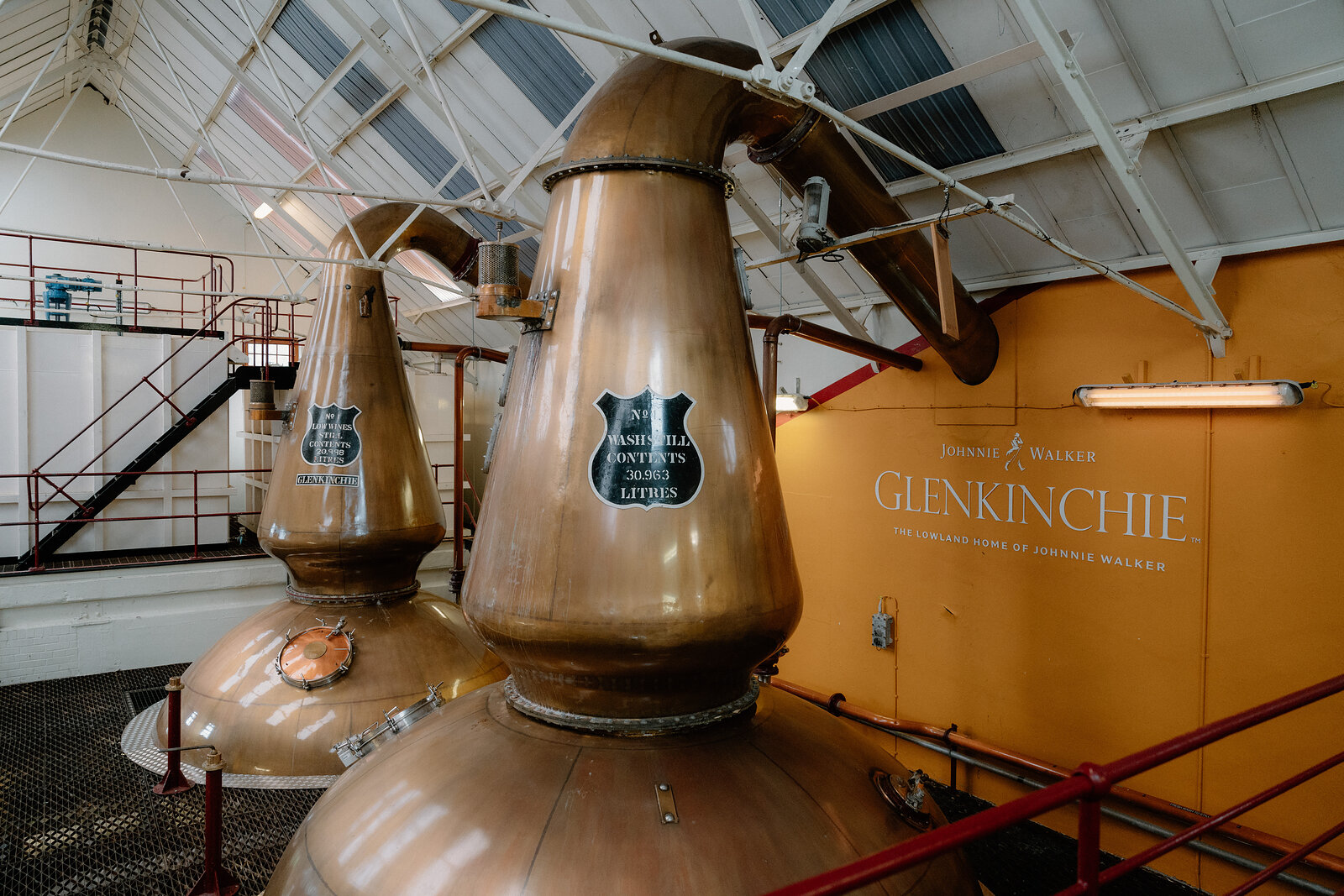 Two copper pot stills stand before an orange wall lettered 'GLENKINCHIE' beneath a glazed roof.