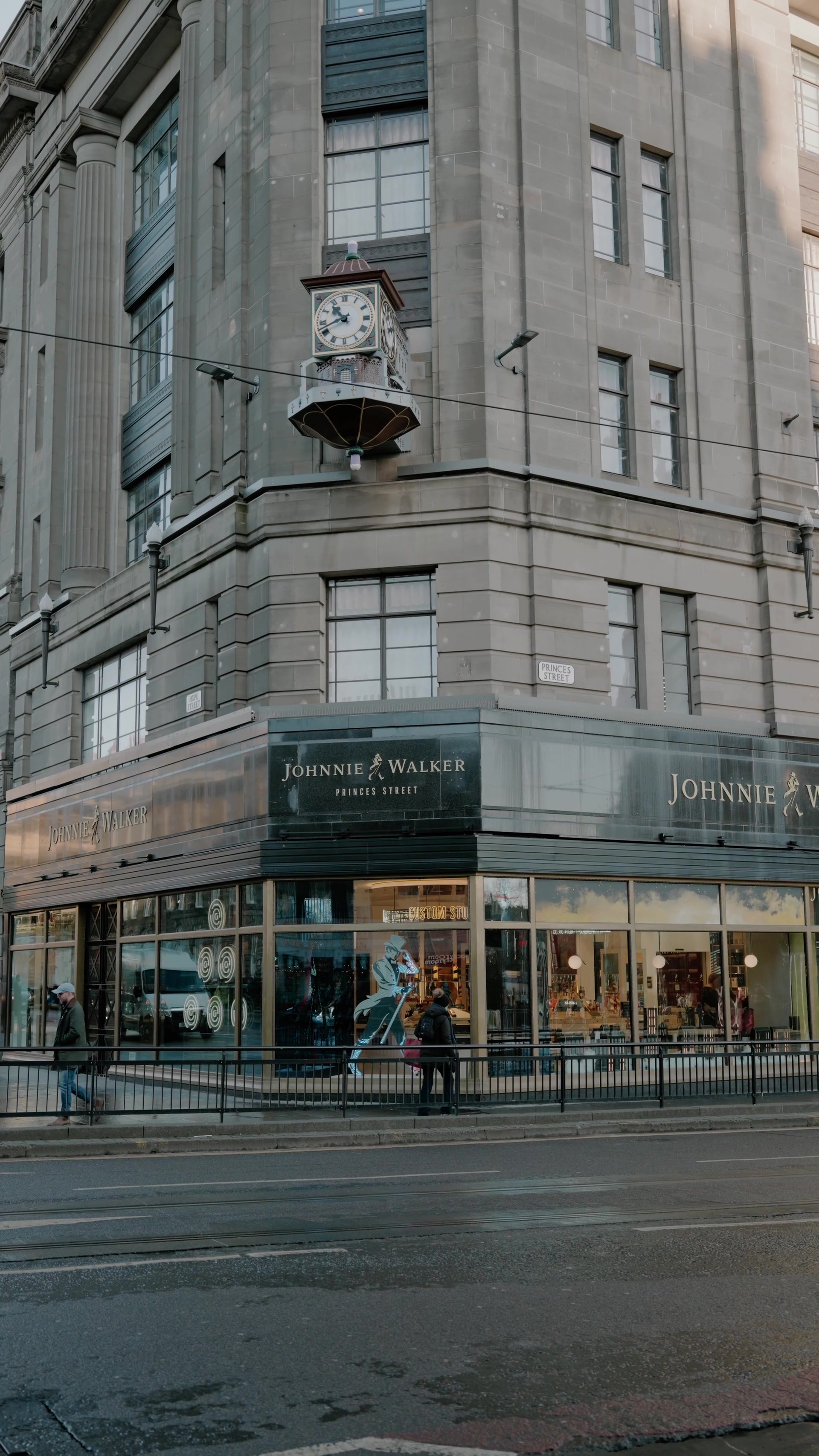 An ornate clock projects from the corner of a sandstone building above the Johnnie Walker Princes Street shopfront in Edinburgh, pedestrians passing below.
