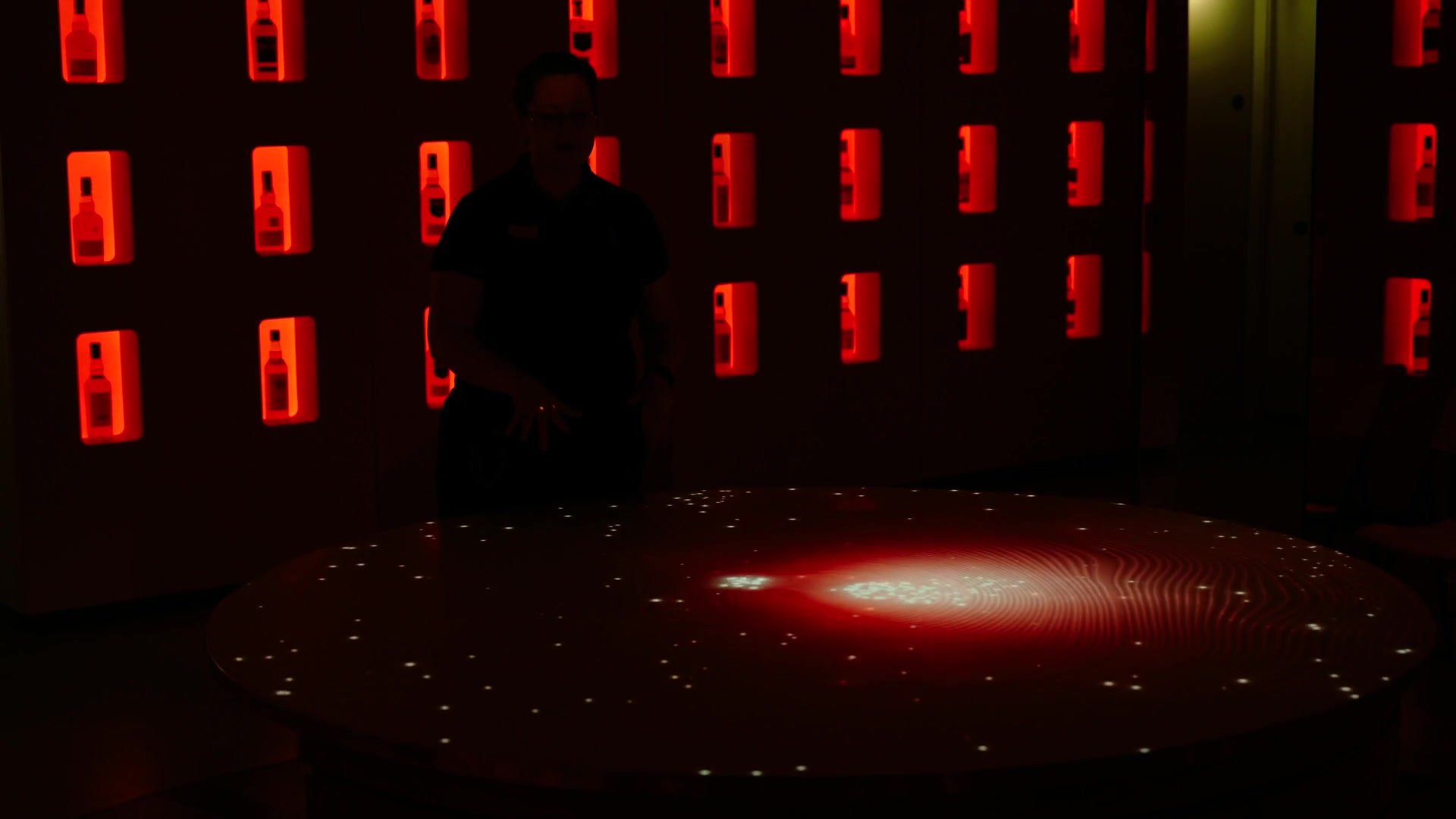 A silhouetted man stands beside a glowing interactive table rippling with light, illuminated whisky bottles set in the wall behind him at the Johnnie Walker Experience.