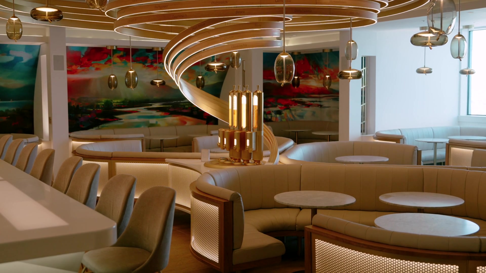 A spiral of wooden distillery-name rings and amber pendant lights above curved banquettes and a gold still sculpture