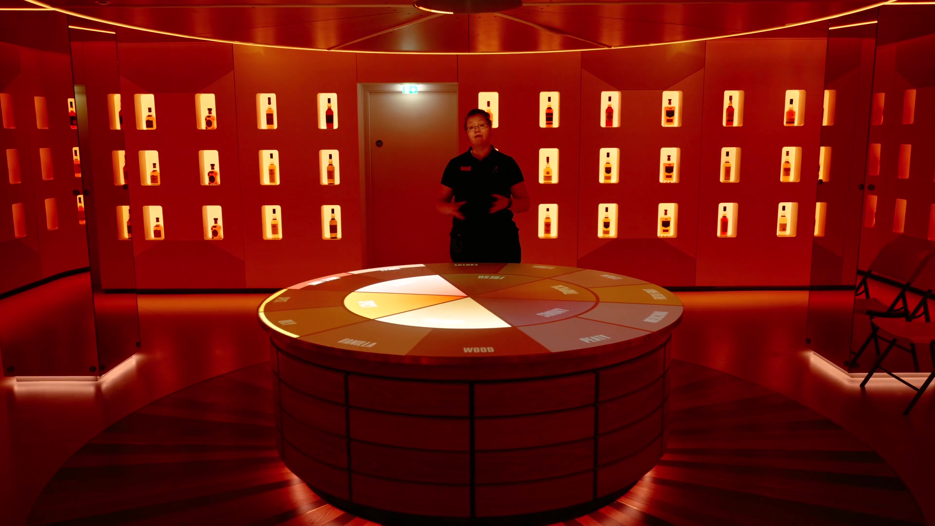 A guide standing beside a circular flavour wheel table lit red beneath rows of illuminated whisky bottles