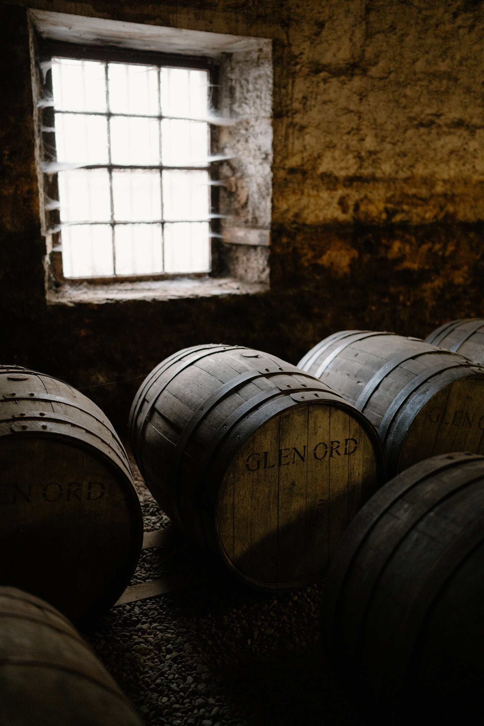Whisky casks branded 'Glen Ord' are stacked beneath a barred window in a warehouse.