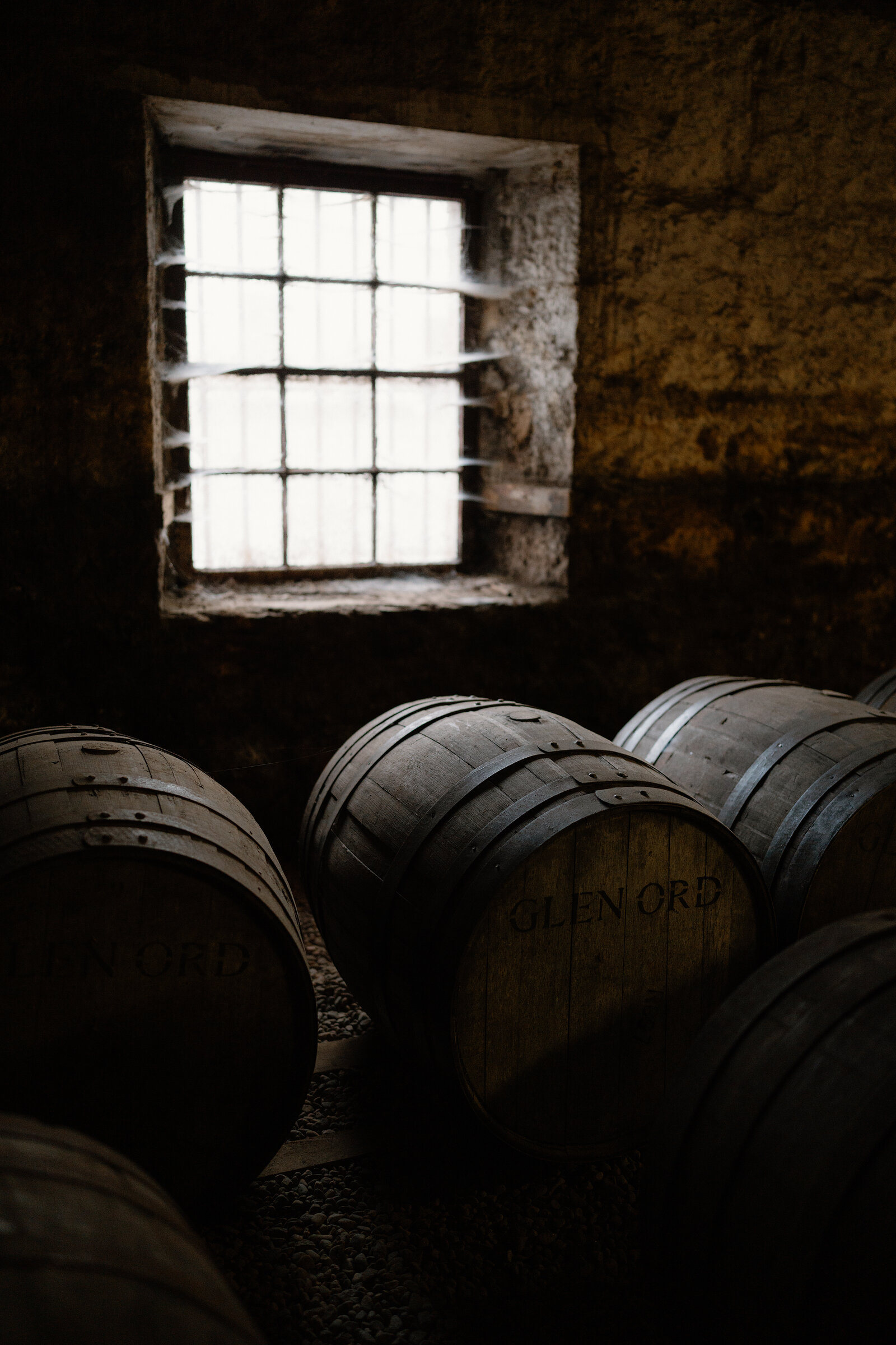 Whisky casks branded 'Glen Ord' are stacked beneath a barred window in a warehouse.