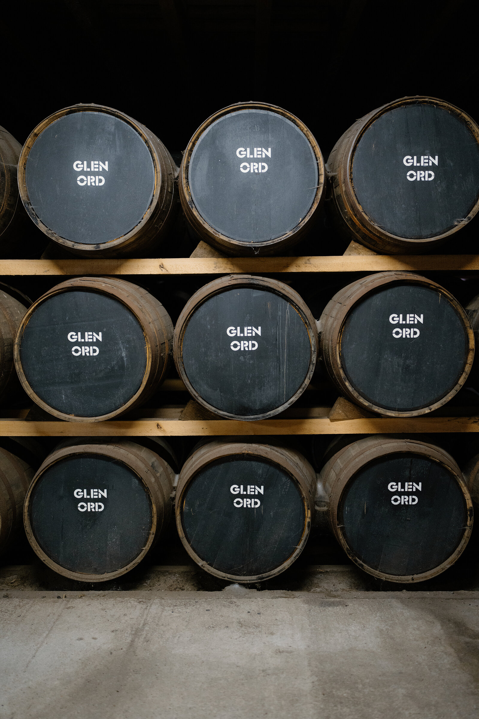 Whisky casks branded 'Glen Ord' are stacked beneath a barred window in a warehouse.
