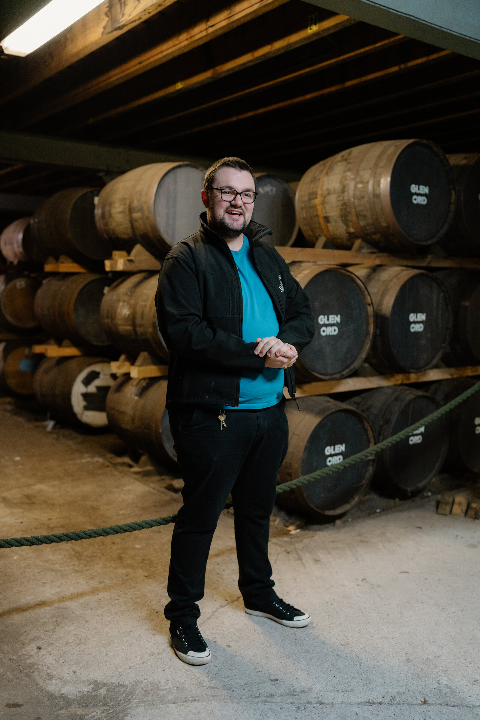 A man stands smiling in front of whisky casks branded 'Glen Ord' in a warehouse.