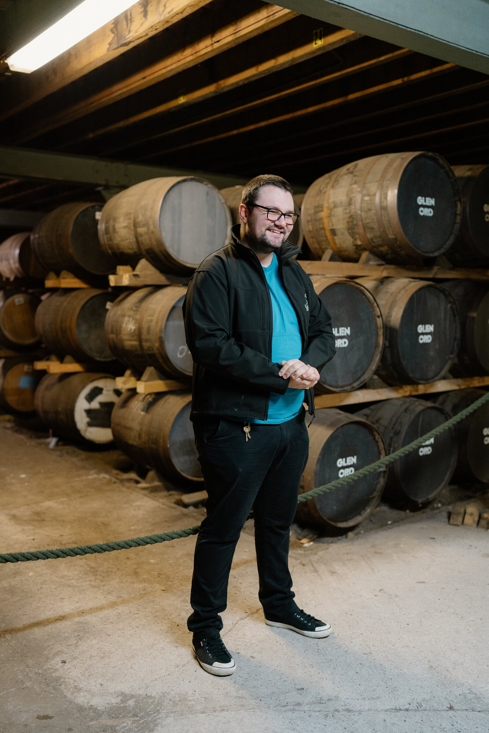 A man stands smiling in front of whisky casks branded 'Glen Ord' in a warehouse.