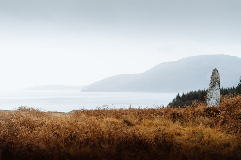 The standing stone of Camus an Staca on a slope of rust-coloured grass, the sea and hills beyond.