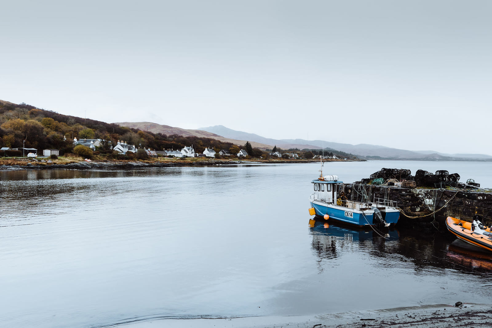A blue fishing boat at Craighouse pier, the village along the bay behind.