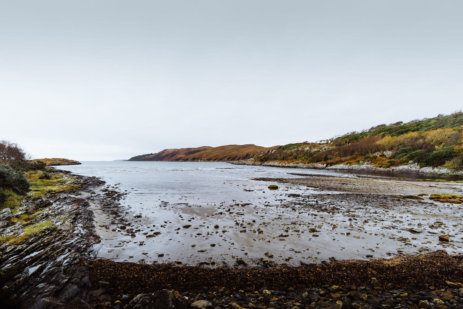 The bay at Inverlussa at low tide, rocks and weed on the sand, hills beyond.
