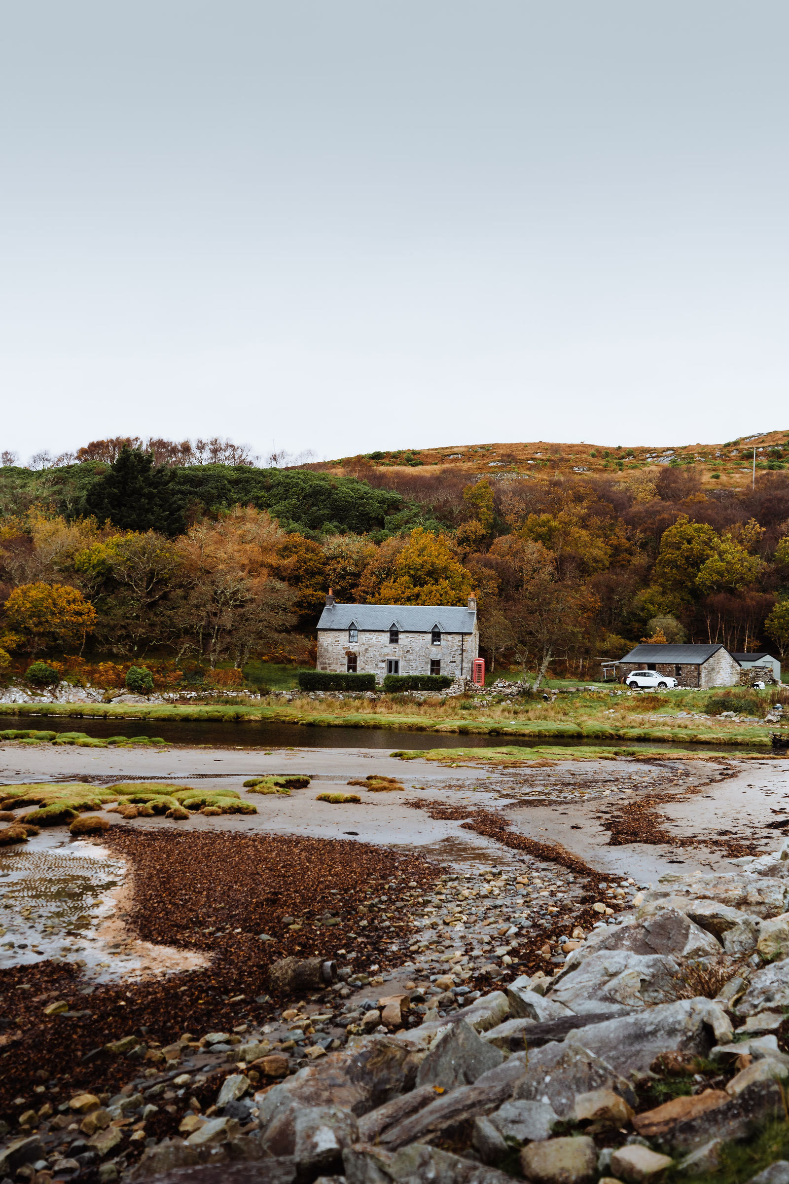 A stone cottage by the tidal shore at Inverlussa, autumn woodland behind.