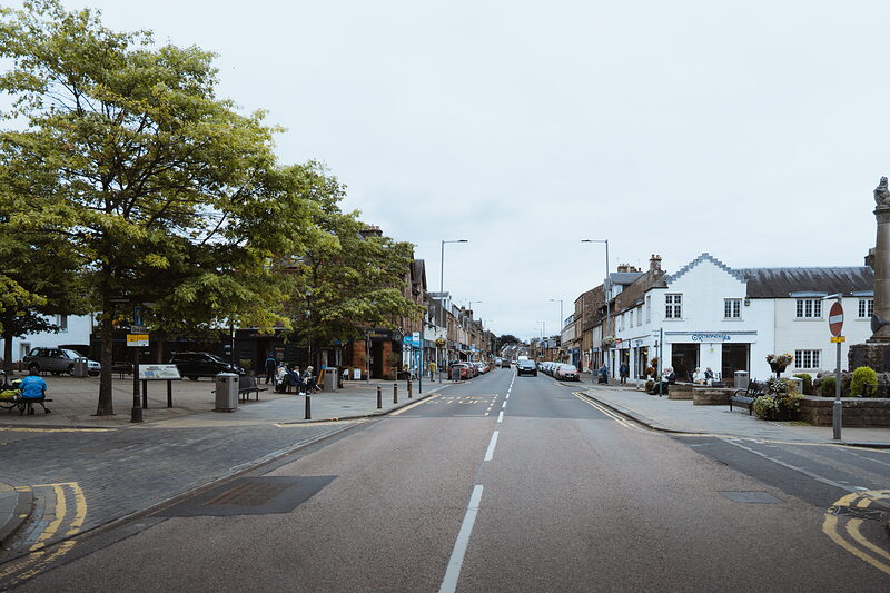 An empty town high street stretches towards a war memorial and church spire, shopfronts and trees lining both sides.
