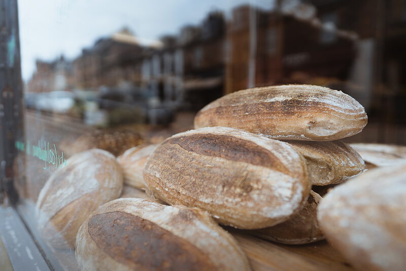 Crusty round loaves are piled in a bakery shop window, a street of buildings reflected faintly in the glass.