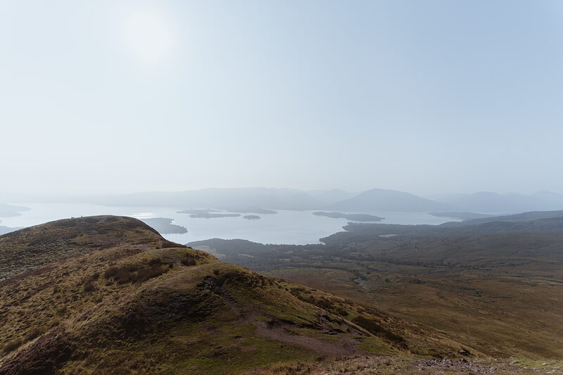 A hazy view from a hilltop looks down over a loch scattered with wooded islands, hills fading into the distance.
