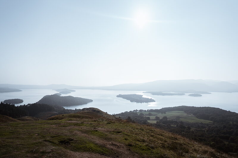 A hazy panoramic view over a loch dotted with wooded islands, the sun glaring low over hills fading into the distance.