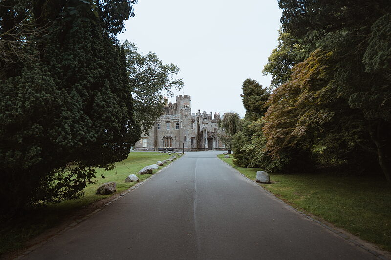 A crenellated stone mansion with towers stands at the end of a long driveway, mature trees lining the approach and lawns either side.