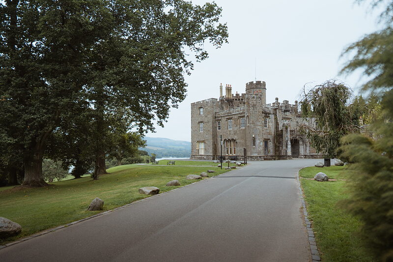 A crenellated stone mansion with corner towers stands beyond parkland trees at the end of a driveway, a glimpse of water visible to one side.