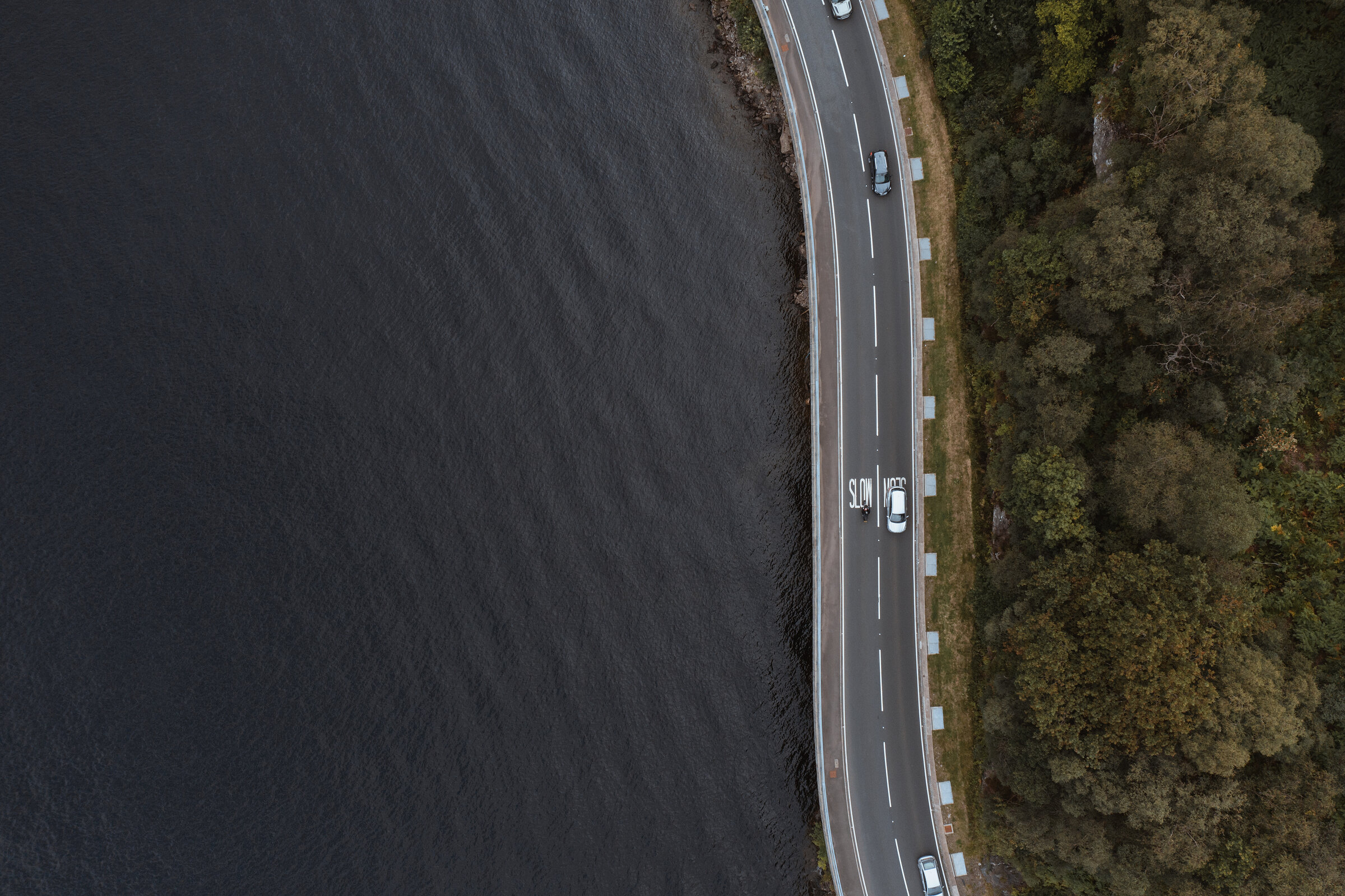 An aerial view looks down a curving road running along a loch shore, cars passing between the water and a bank of autumn-coloured trees.