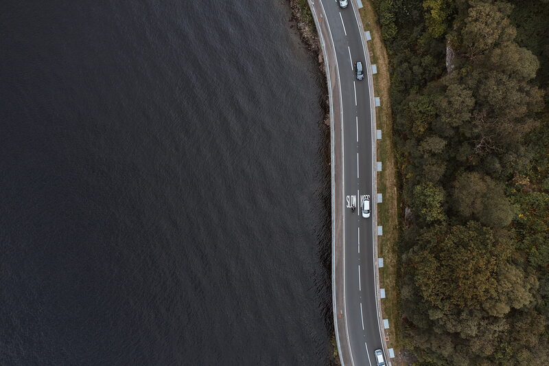 An aerial view looks down a curving road running along a loch shore, cars passing between the water and a bank of autumn-coloured trees.