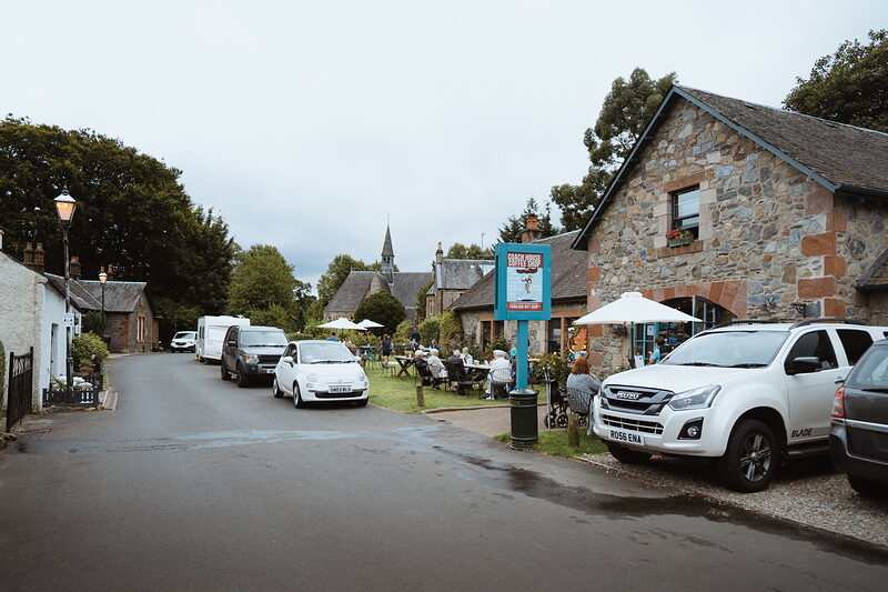 Cars line a village street past a café with tables under a parasol, a church spire rising above the rooftops beyond.