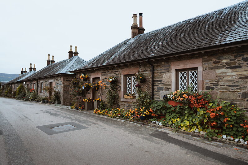 A row of stone cottages with tall chimneys is fronted by nasturtiums and hanging flower baskets, a quiet road running past.