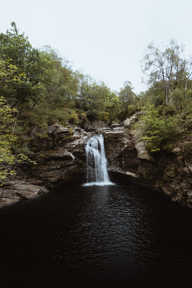 A waterfall drops into a dark rock pool within a wooded gorge, moss-covered boulders framing the falling water.