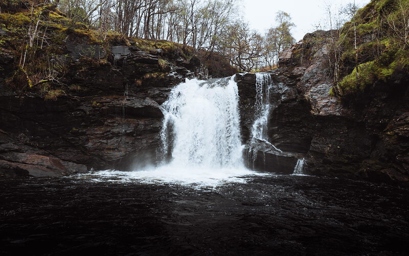 A waterfall drops into a dark pool between rocky, moss-covered cliffs, bare trees rising along the top of the gorge.