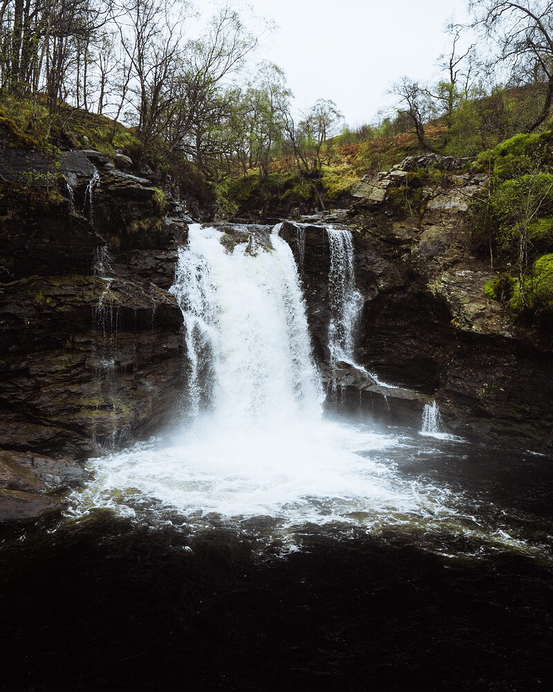 A waterfall drops over dark rock ledges into a pool, bare trees and moss-covered banks framing the scene.