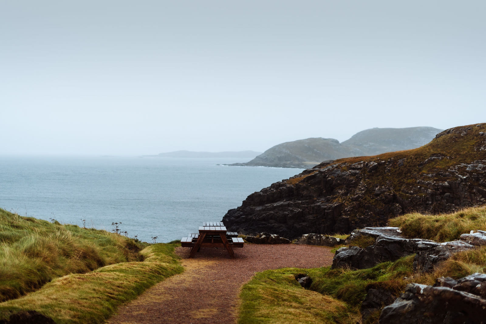A picnic table at the end of a gravel path on the headland, dark rocks and grassy slopes falling to the sea and a hilly coast beyond.