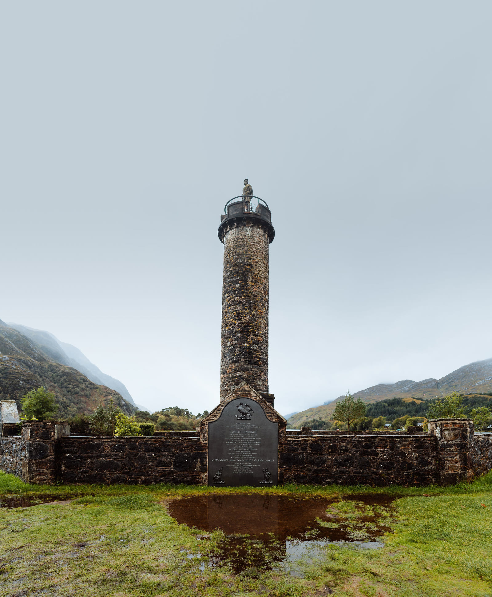 The Glenfinnan Monument inside its walled enclosure, a puddle reflecting the tower.