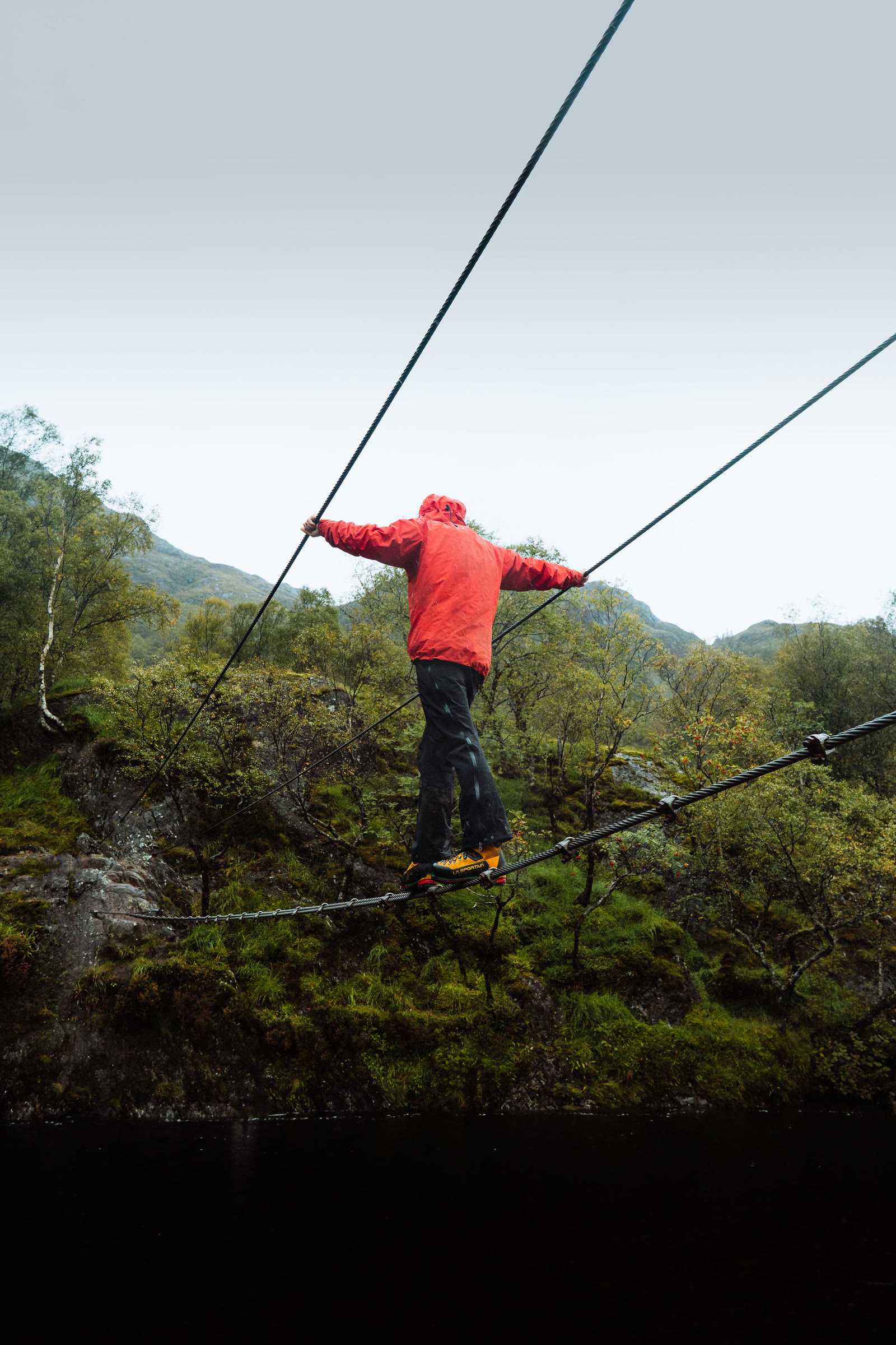 A walker in a red jacket halfway across the wire bridge at Steall, arms out on the two hand wires.